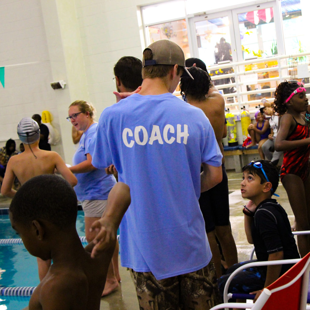 A swim coach is talking with his teammates on the pool deck during a swim meet. The swimmer is waiting in line for his turn to race.