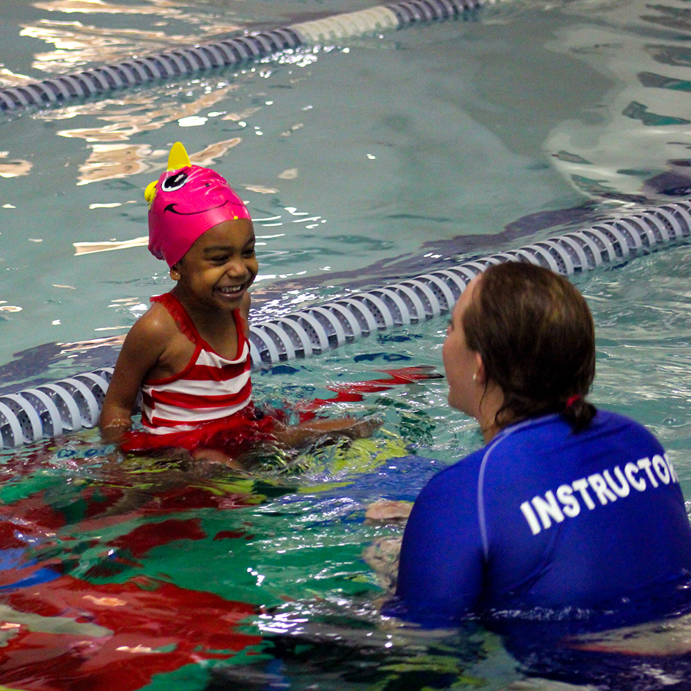 A young swim student is sitting on a platform in the water and smiling at her swim instructor who is with arm's reach in the pool and is smiling back at the swimmer.