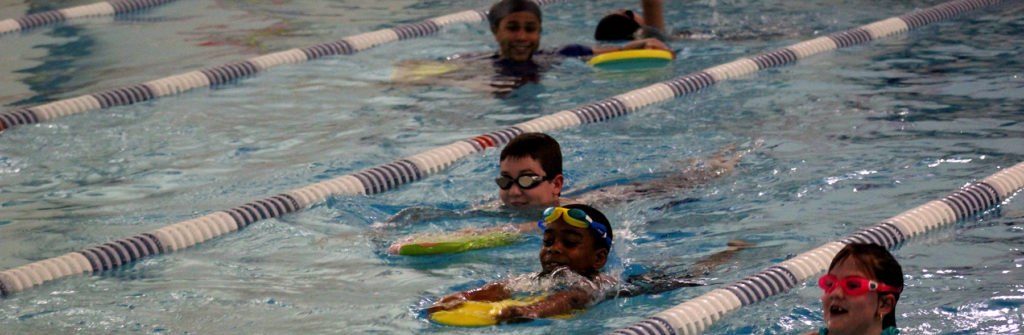 A group of swim students are using kickboards to swim laps in the pool. The swimmer in front has his goggles up and is smiling. Behind the three students, their swim instructor is watching and smiling.