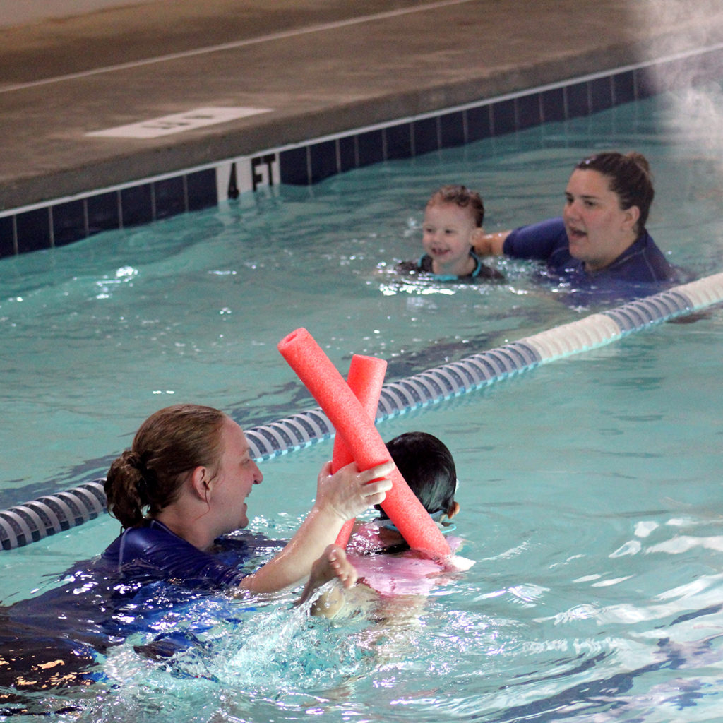 Two swim instructors are aiding their swim students in practicing swim lessons. Both instructors have 1 student and are holding on to them to support them above the water.