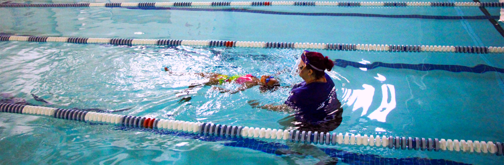 An instructor is in the water with a student. The instructor is walking backwards with the student swimming up towards her. The student is swimming on her back and kicking up the lane towards the instructor.